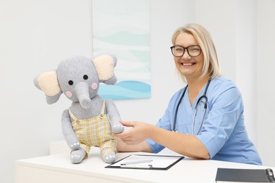 Professional doctor with clipboard and toy elephant at white table in clinic