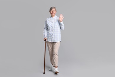 Senior woman with walking cane waving on gray background