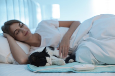 Photo of Young woman and cat in bed with electric heating pad, focus on cable