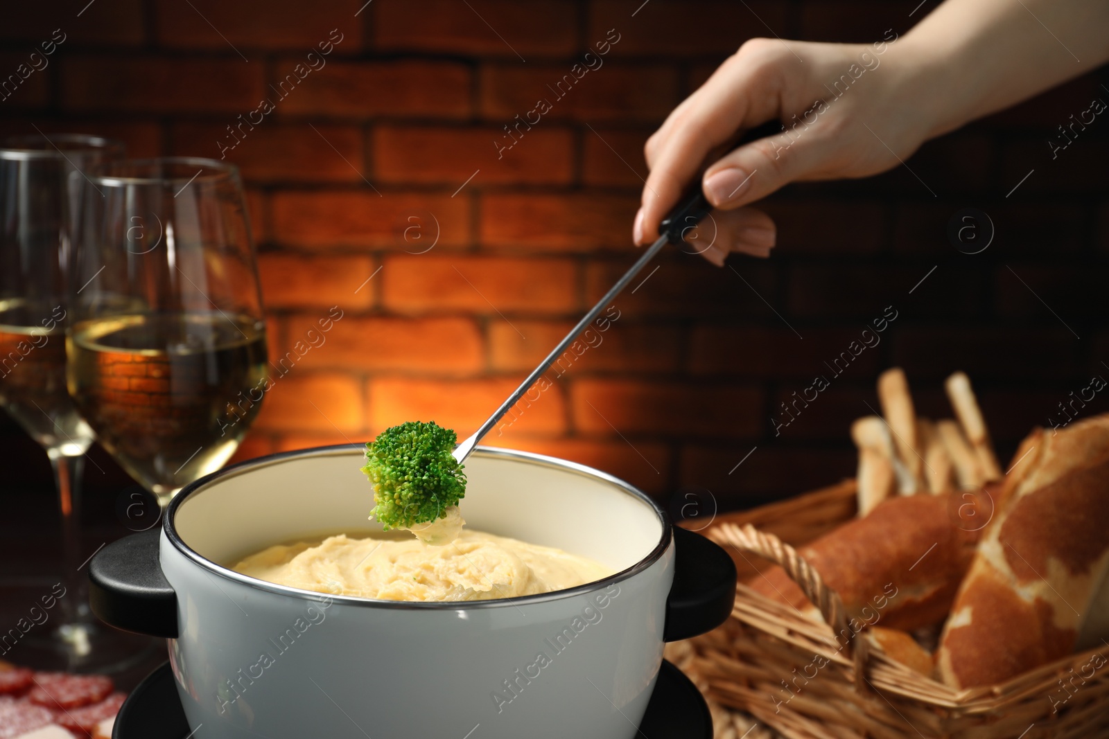 Photo of Woman dipping piece of broccoli into fondue pot with melted cheese at wooden table with wine and snacks, closeup