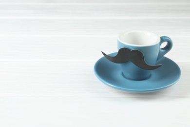 Photo of Cup with fake paper mustache and saucer on white wooden table, space for text