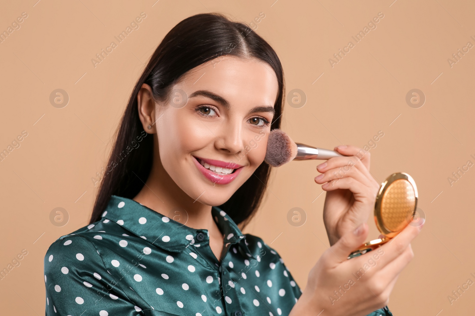 Photo of Happy woman with cosmetic pocket mirror applying makeup on light brown background