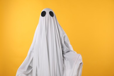 Photo of Creepy ghost. Person covered with white sheet on yellow background, space for text