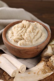 Photo of Spicy horseradish sauce in bowl and roots on table, closeup