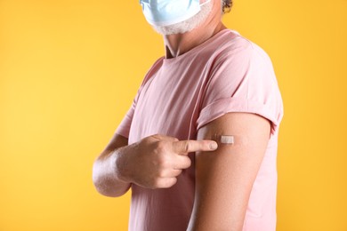 Photo of Senior man in protective mask pointing at arm with bandage after vaccination on yellow background, closeup