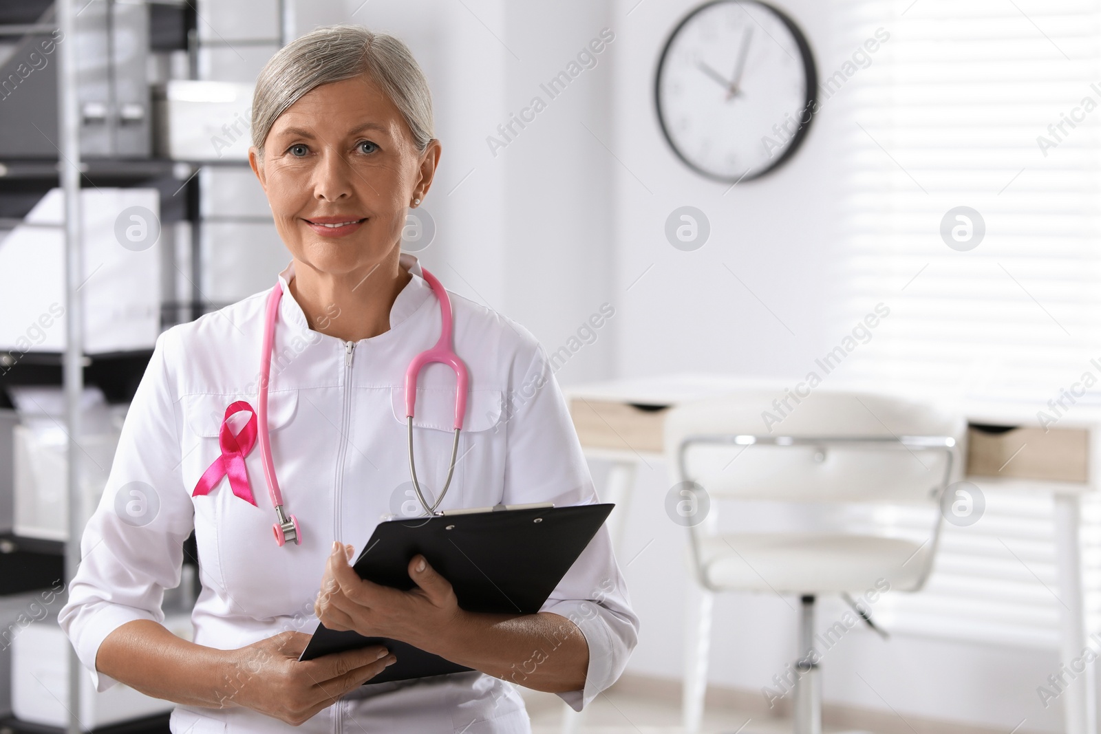 Photo of Doctor with stethoscope, clipboard and pink ribbon indoors, space for text. Breast cancer awareness