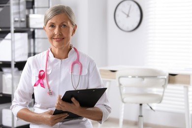 Photo of Doctor with stethoscope, clipboard and pink ribbon indoors, space for text. Breast cancer awareness