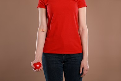 Photo of Blood donation concept. Woman with adhesive plaster on arm holding red heart against brown background, closeup. Space for text