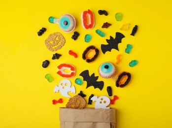 Paper bag of tasty candies and Halloween decorations on yellow background, flat lay