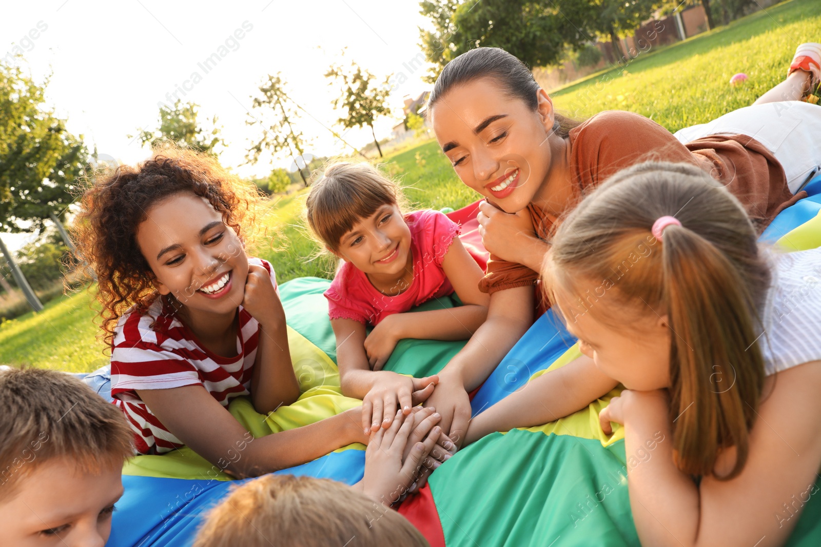 Photo of Group of children with teachers holding hands together on rainbow playground parachute in park. Summer camp activity