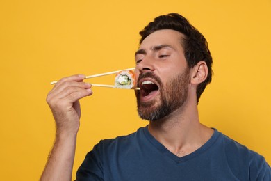 Handsome man eating tasty sushi roll with chopsticks on orange background