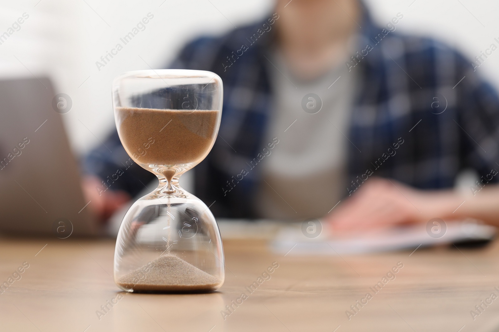 Photo of Hourglass with flowing sand on desk. Man taking notes while using laptop indoors, selective focus