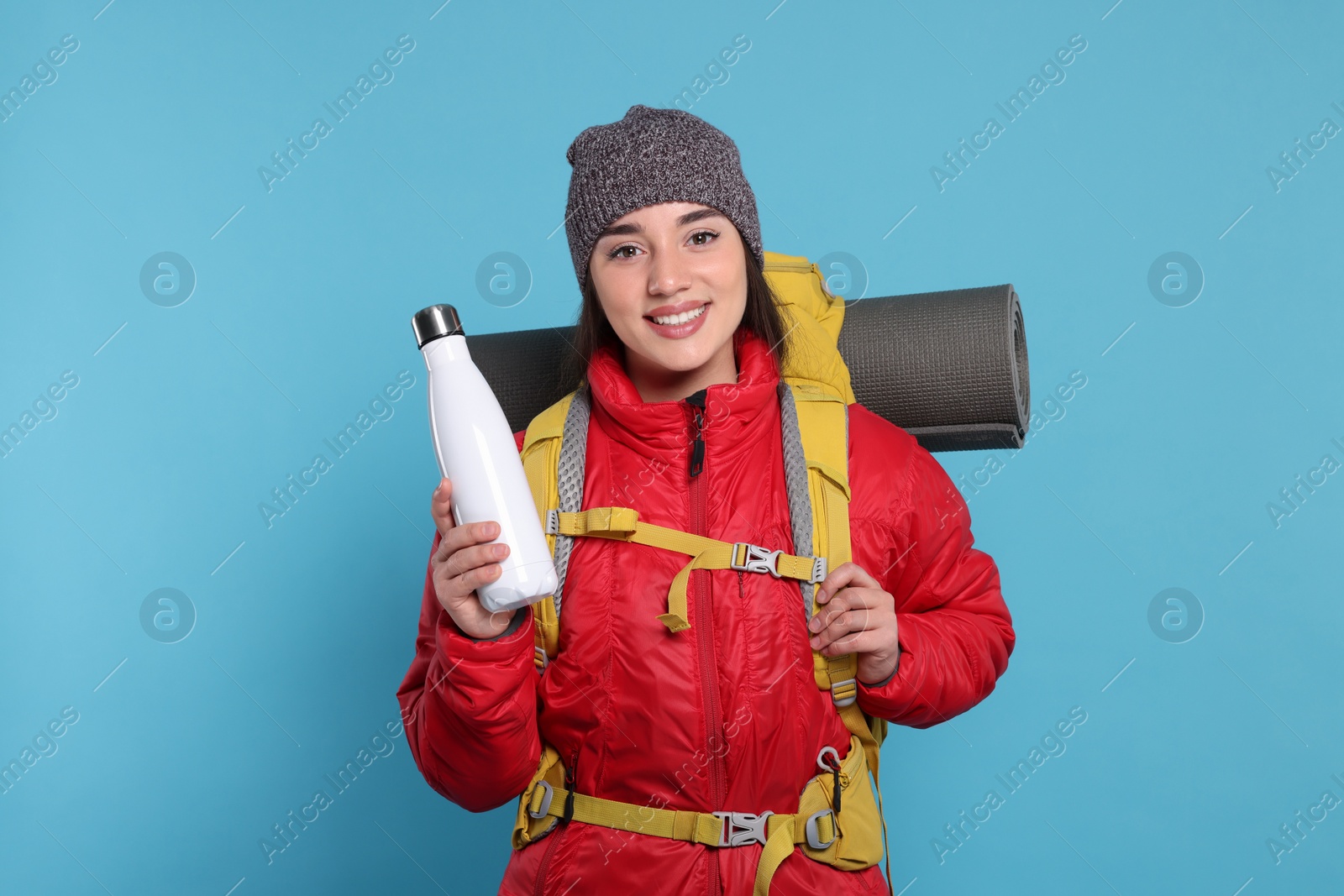 Photo of Smiling young woman with backpack and thermo bottle on light blue background. Active tourism