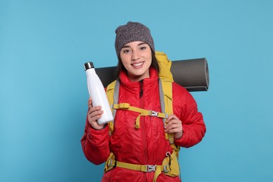 Photo of Smiling young woman with backpack and thermo bottle on light blue background. Active tourism