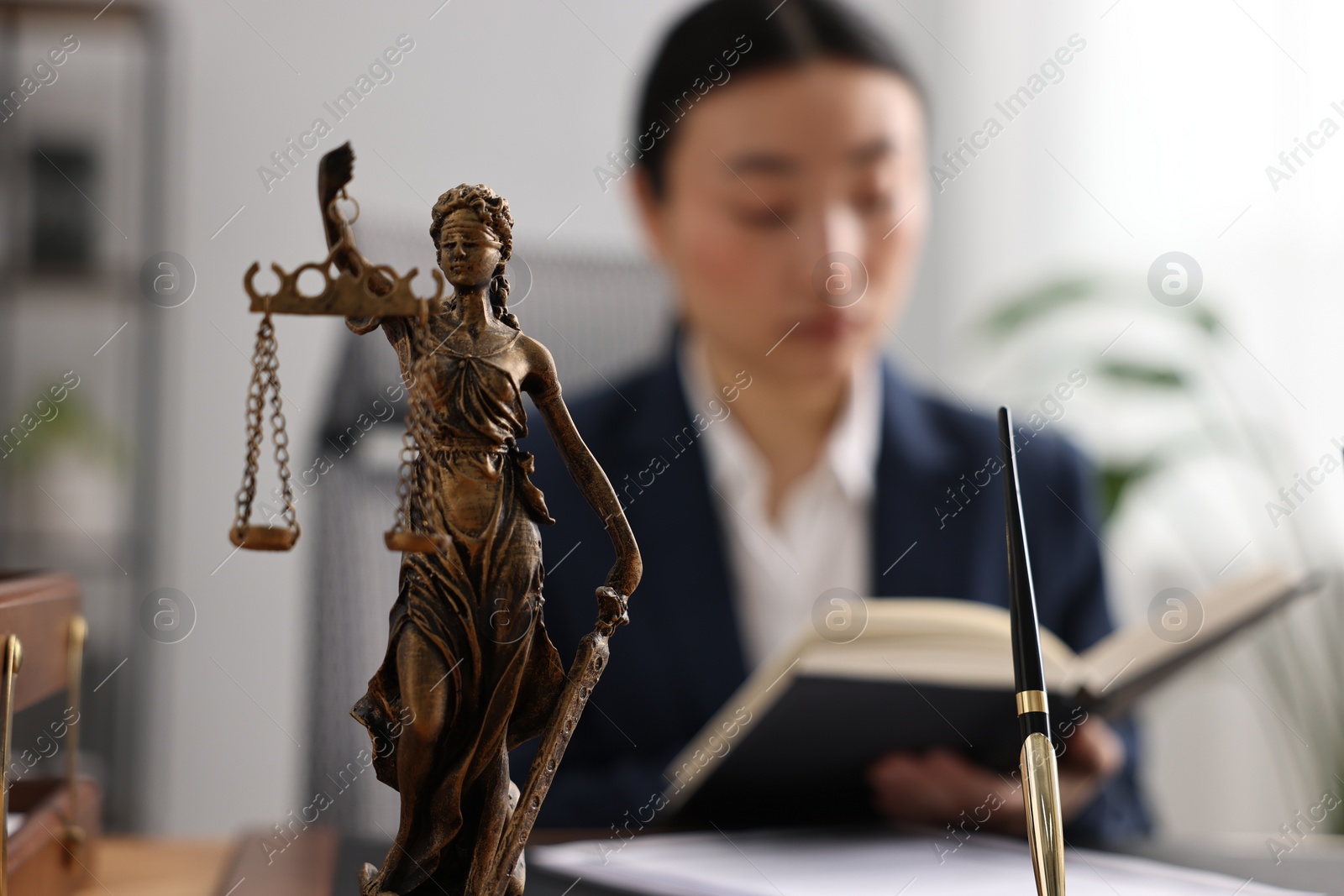 Photo of Notary reading book at table in office, focus on statue of Lady Justice and pen