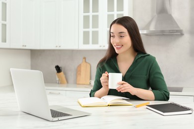 Photo of Home workplace. Happy woman with cup of hot drink looking at laptop at marble desk in kitchen