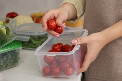 Woman putting cherry tomatoes into container with fresh vegetables at white marble table in kitchen, closeup. Food storage