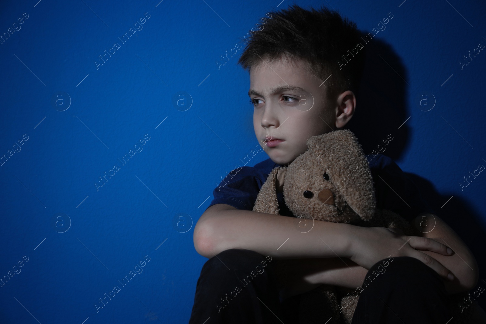 Photo of Sad little boy with toy near blue wall, space for text. Domestic violence concept