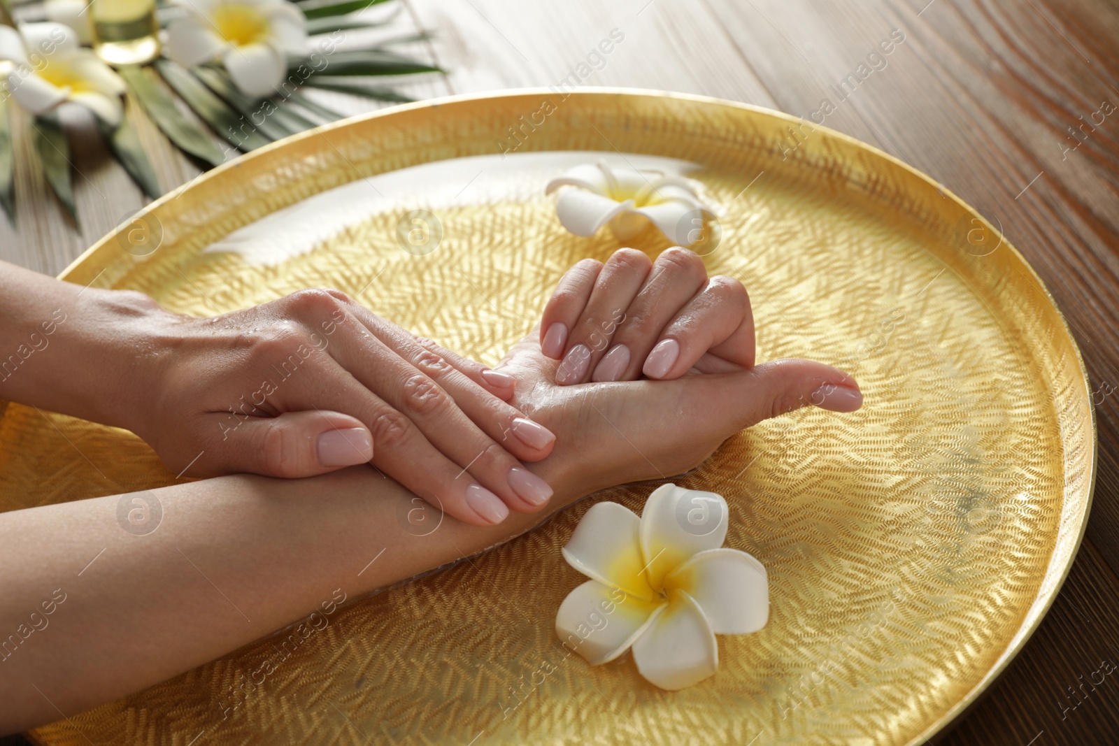 Photo of Woman soaking her hands in bowl with water and flowers on table, closeup. Spa treatment