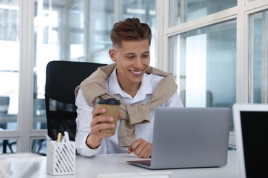Happy man with paper cup of coffee working on laptop in office
