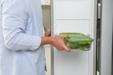 Photo of Man putting container with fresh broccoli into fridge in kitchen, closeup. Food storage