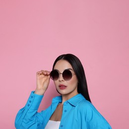 Attractive serious woman touching fashionable sunglasses against pink background. Space for text