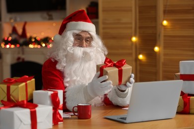 Photo of Santa Claus with Christmas gift using laptop at table in room