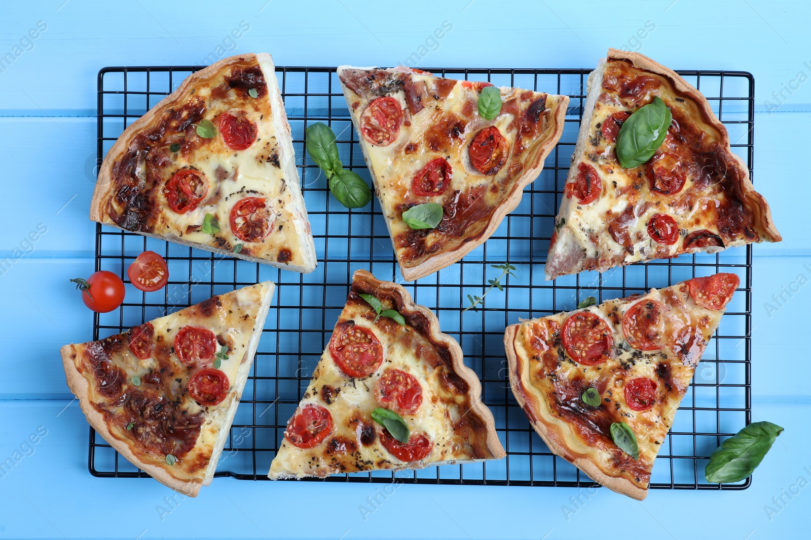 Photo of Pieces of delicious homemade quiche with prosciutto, tomatoes and basil on light blue wooden table, flat lay