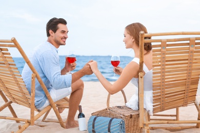 Happy young couple with glasses of wine sitting on deck chairs at sea beach