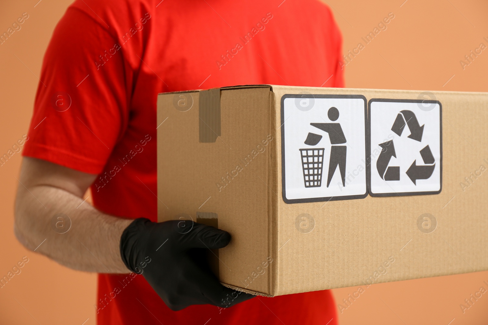 Photo of Courier holding cardboard box with different packaging symbols on orange background, closeup. Parcel delivery