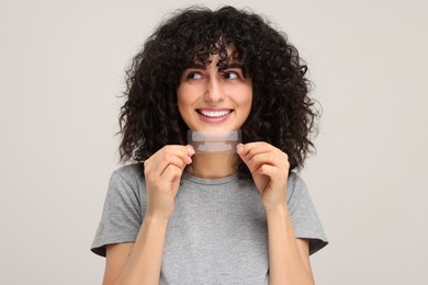 Young woman holding teeth whitening strips on light grey background