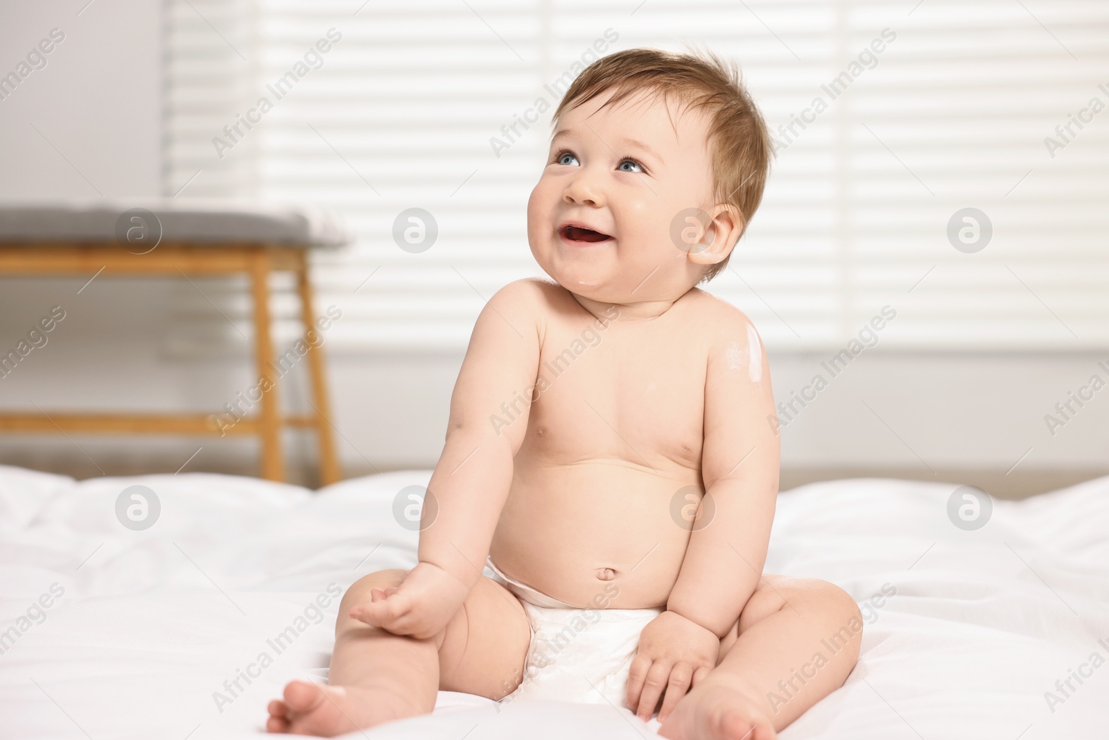Photo of Cute little baby with moisturizing cream on body sitting on bed at home