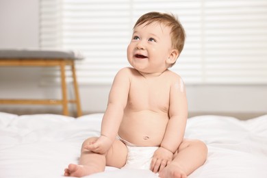 Cute little baby with moisturizing cream on body sitting on bed at home