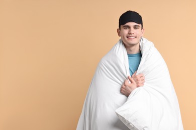 Photo of Happy man in pyjama and sleep mask wrapped in blanket on beige background, space for text