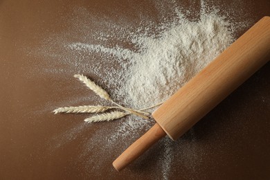 Photo of Flour, spikelets and rolling pin on brown table, top view