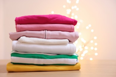 Photo of Stack of folded clothes on wooden table against beige background with blurred lights, space for text