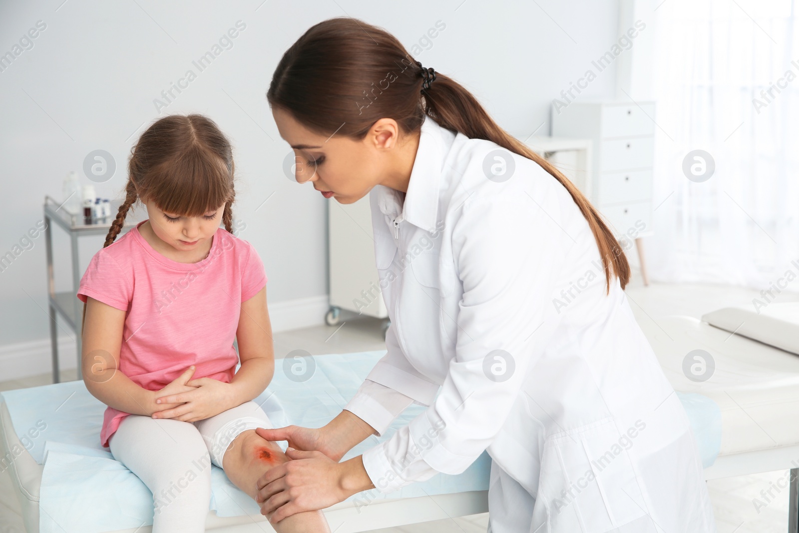 Photo of Female doctor examining little girl's injured leg in clinic. First aid