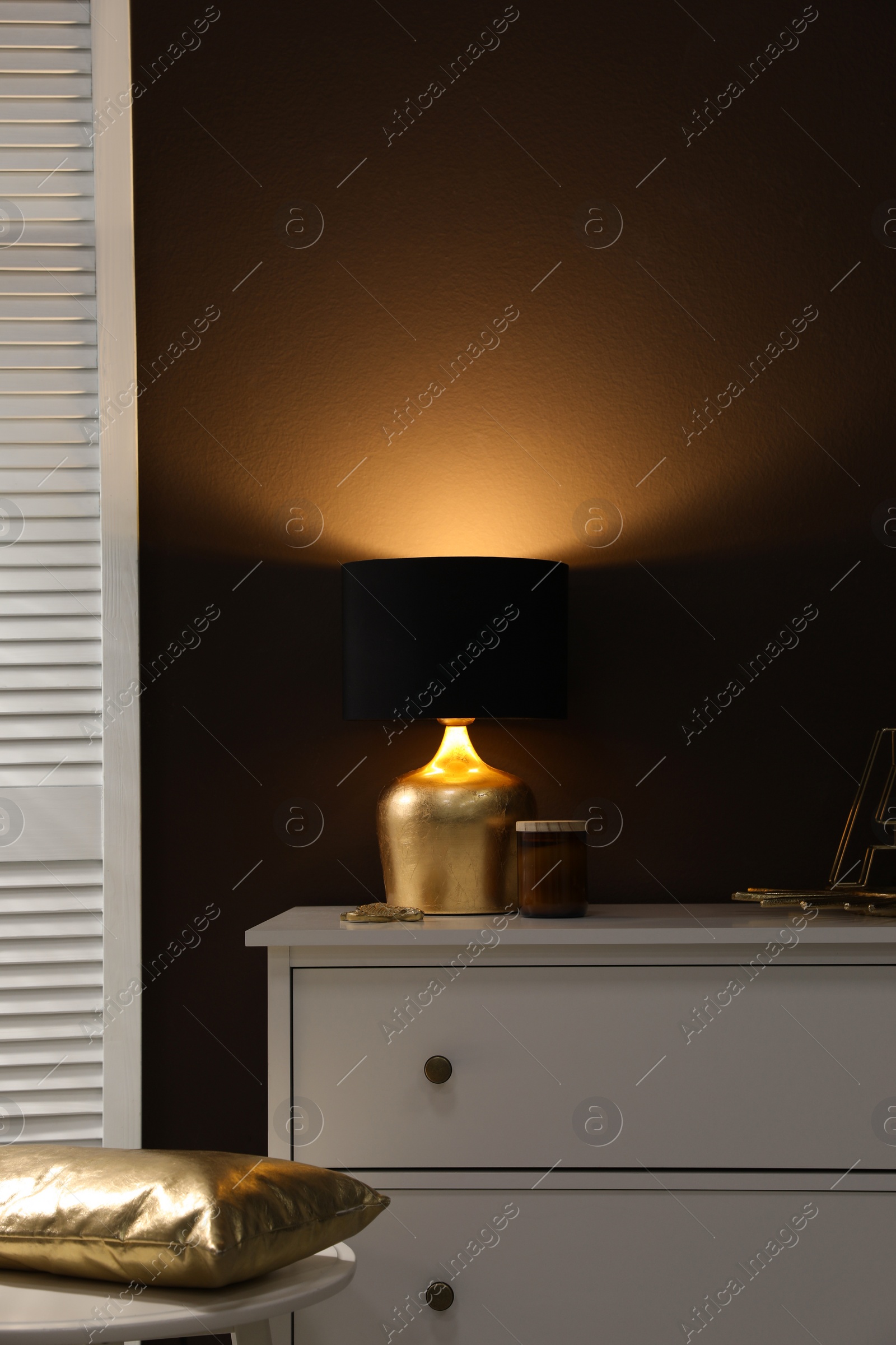 Photo of Stylish lamp and decor on white chest of drawers near brown wall indoors. Interior design