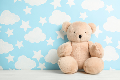 Photo of Teddy bear on white wooden table near wall with painted blue sky, space for text. Baby room interior
