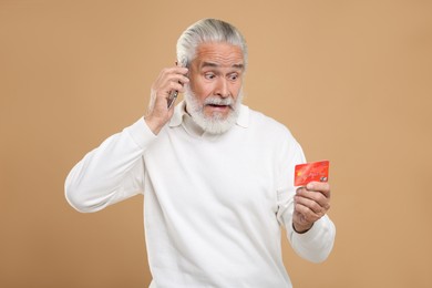 Photo of Shocked senior man with credit card talking on smartphone against beige background. Be careful - fraud