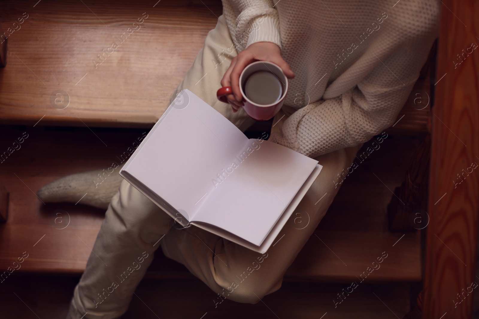 Photo of Woman with cup of coffee reading book on stairs at home, top view