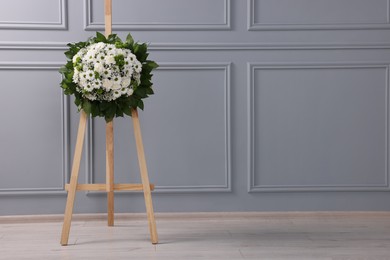 Funeral wreath of flowers on wooden stand near light grey wall indoors, space for text