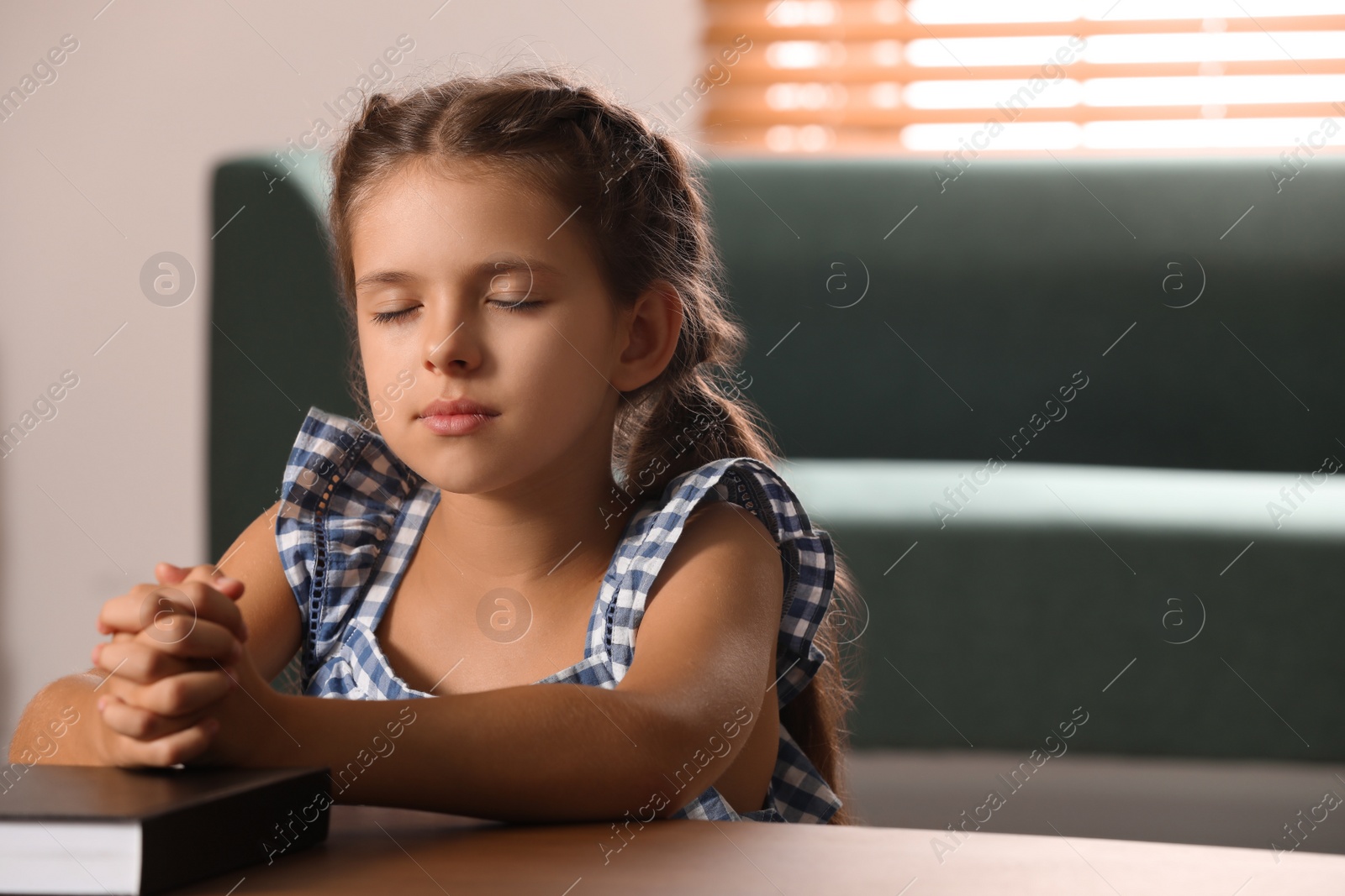 Photo of Cute little girl praying over Bible at table in room. Space for text