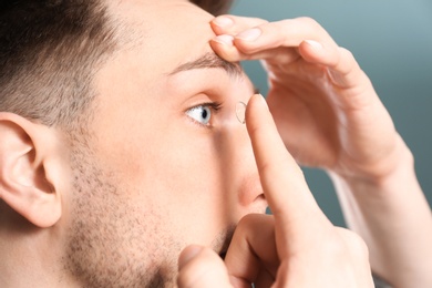 Young man putting contact lens in his eye on color background