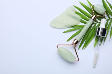 Photo of Gua sha stone, face roller, dropper and green leaves on white background, flat lay. Space for text