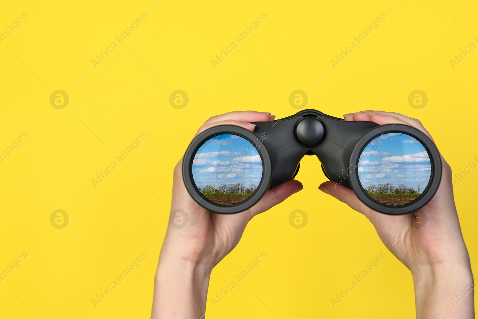 Image of Woman holding binoculars on yellow background, closeup. Country landscape reflecting in lenses