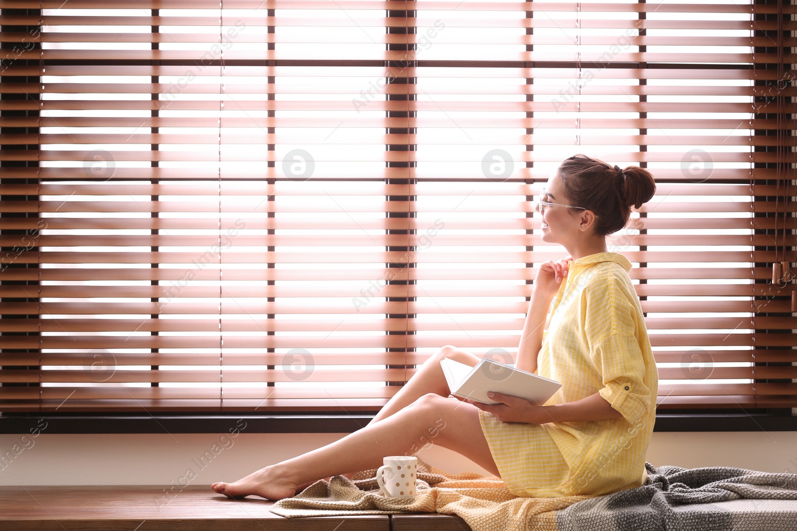 Photo of Young woman reading book near window with blinds at home. Space for text