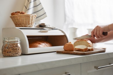 Photo of Woman cutting loaf near wooden bread basket at white marble table in kitchen, closeup
