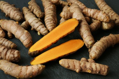Photo of Whole and cut turmeric roots on black textured table, closeup