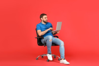 Young man with laptop sitting in comfortable office chair on red background